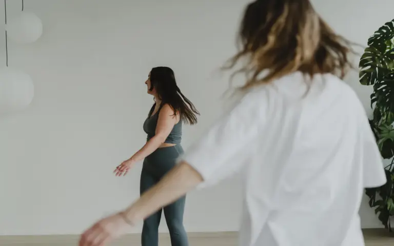Osallistuja liikkuu vapaasti valkoisessa paidassa, hiukset heilahtaen, ja taustalla ohjaaja liikkuu valoisalla salilla barajoogan studiossa sompasaaressa
