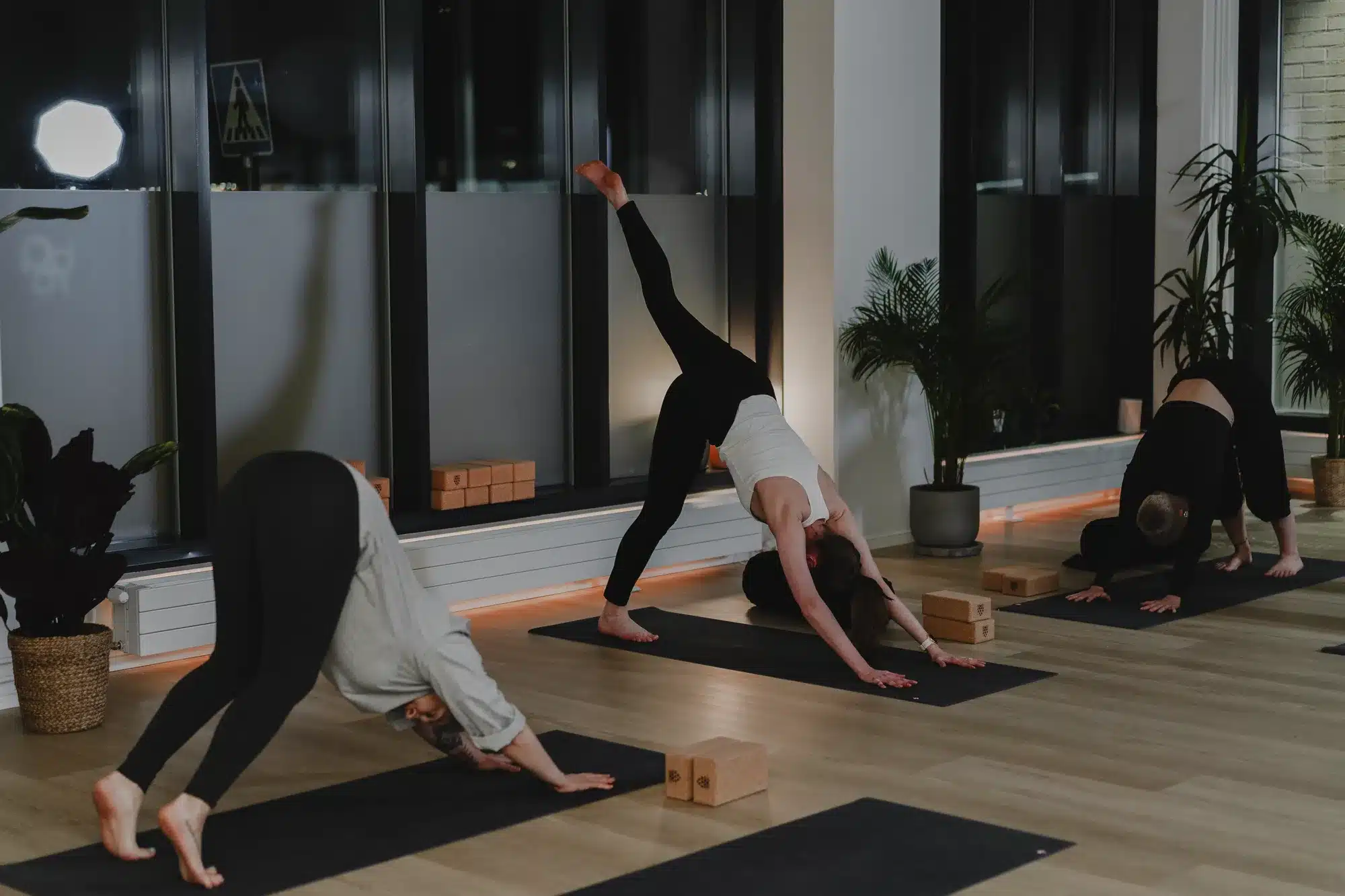 Kolme osallistujaa tekee vinyasa flow -tunnilla alaspäin katsovan koiran asentoa, yksi jalka ilmassa, valoisalla joogasalilla barajoogan studiossa sompasaaressa